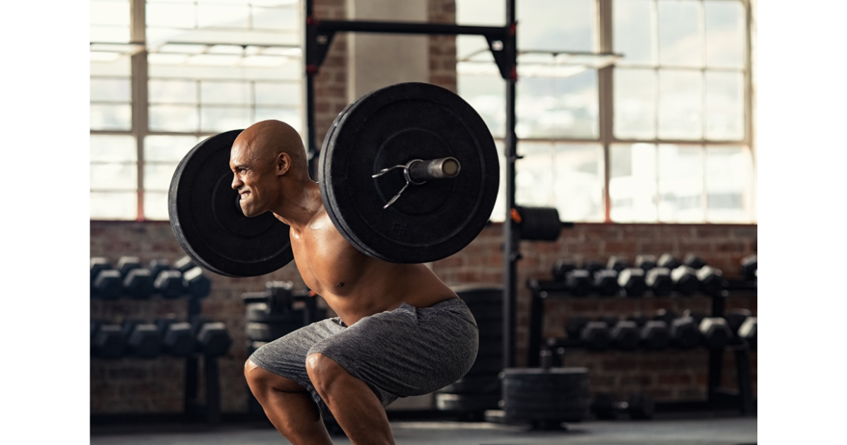 Male athlete performing a heavy barbell back squat in a warehouse-style gym, showing high effort with free weights and a power rack in the background